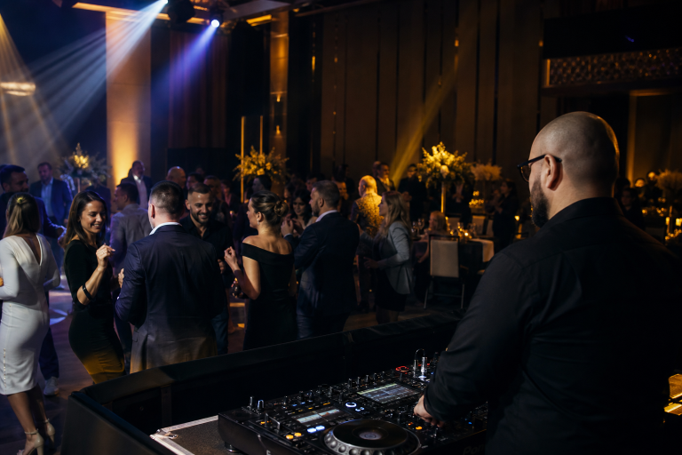 Invitați de vârste diferite intră treptat pe ring la o petrecere corporate, în timp ce DJ-ul urmărește atmosfera din sală.
